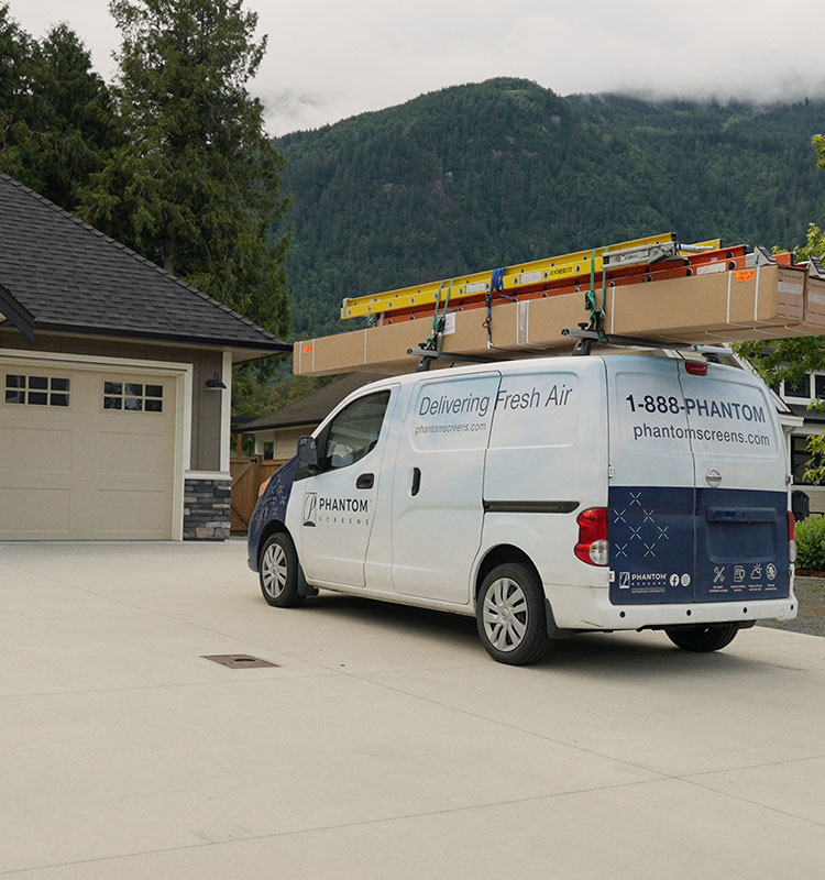 Fourgonnette de service Phantom Screens avec échelles sur le toit, garée dans l'allée d'une maison beige, montagnes en arrière-plan