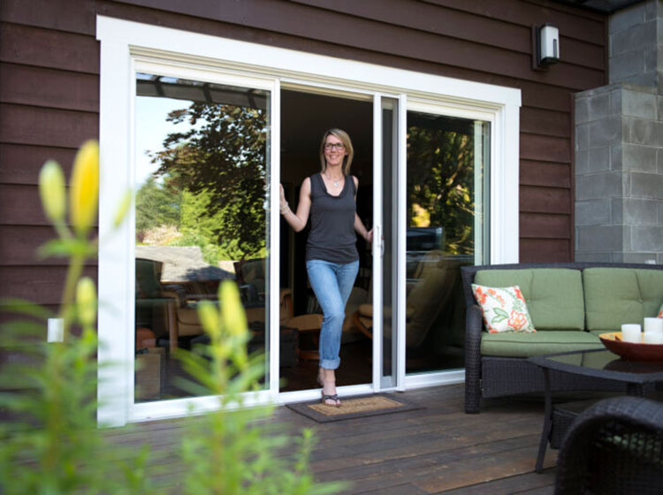 Femme debout dans une porte vitrée coulissante avec moustiquaire rétractable, patio extérieur avec mobilier visible à travers la porte