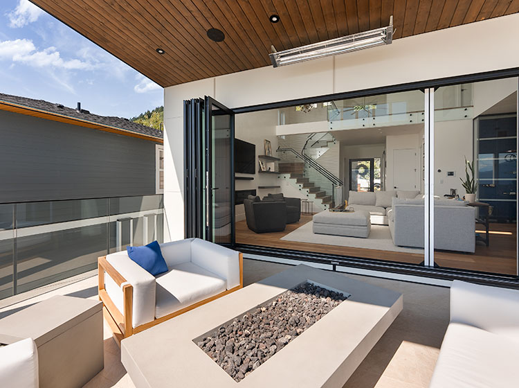Patio moderne avec système de mur vitré pliant entièrement ouvert sur un salon luxueux, avec sièges d'extérieur blancs et foyer intégré