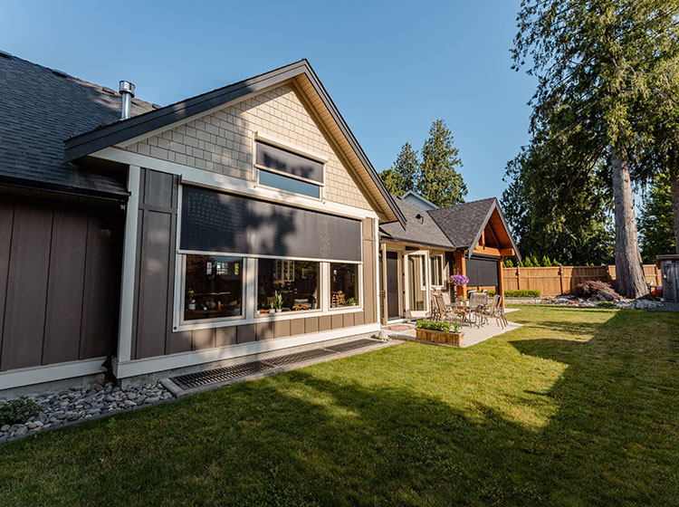 Extérieur de maison artisanale moderne avec stores solaires foncés sur les fenêtres, présentant un jardin paysager et un espace de détente extérieur