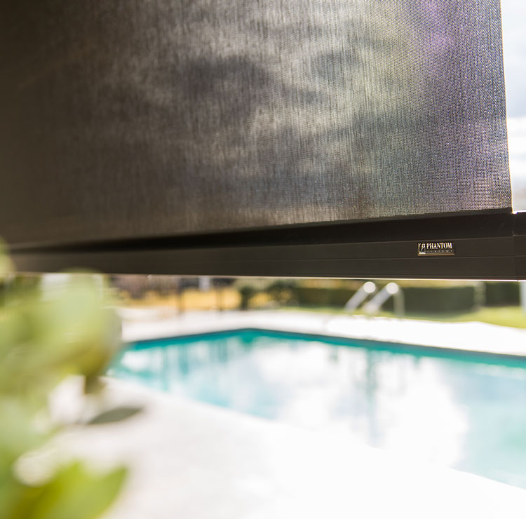 Store solaire extérieur partiellement baissé avec système de guide-câble visible, donnant sur une piscine et des meubles de jardin