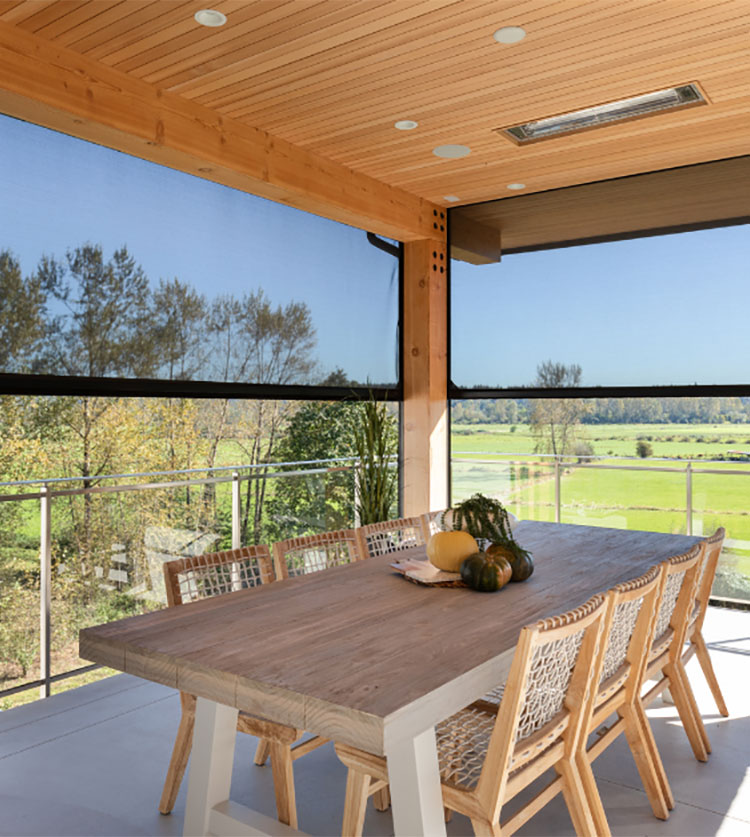 Terrasse couverte avec une table à manger en bois, des chaises en osier et des moustiquaires rétractables motorisées donnant sur des champs verdoyants.