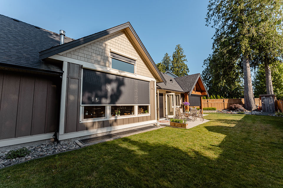 Maison moderne avec moustiquaires rétractables extérieures installées sur les fenêtres, avec jardin paysager, coin terrasse et grands arbres.