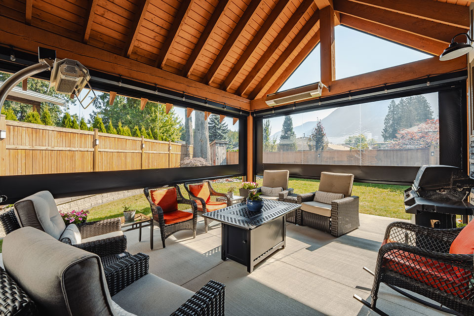 Terrasse couverte avec plafond à poutres en bois et moustiquaires rétractables motorisées, avec mobilier d'extérieur confortable, une table-foyer et vue sur les montagnes en arrière-plan.